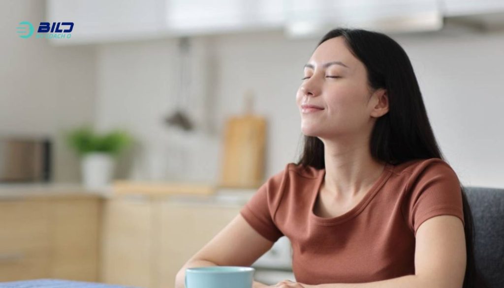 A relaxed woman with closed eyes and a gentle smile sitting in a bright kitchen, enjoying a morning beverage from a blue mug.