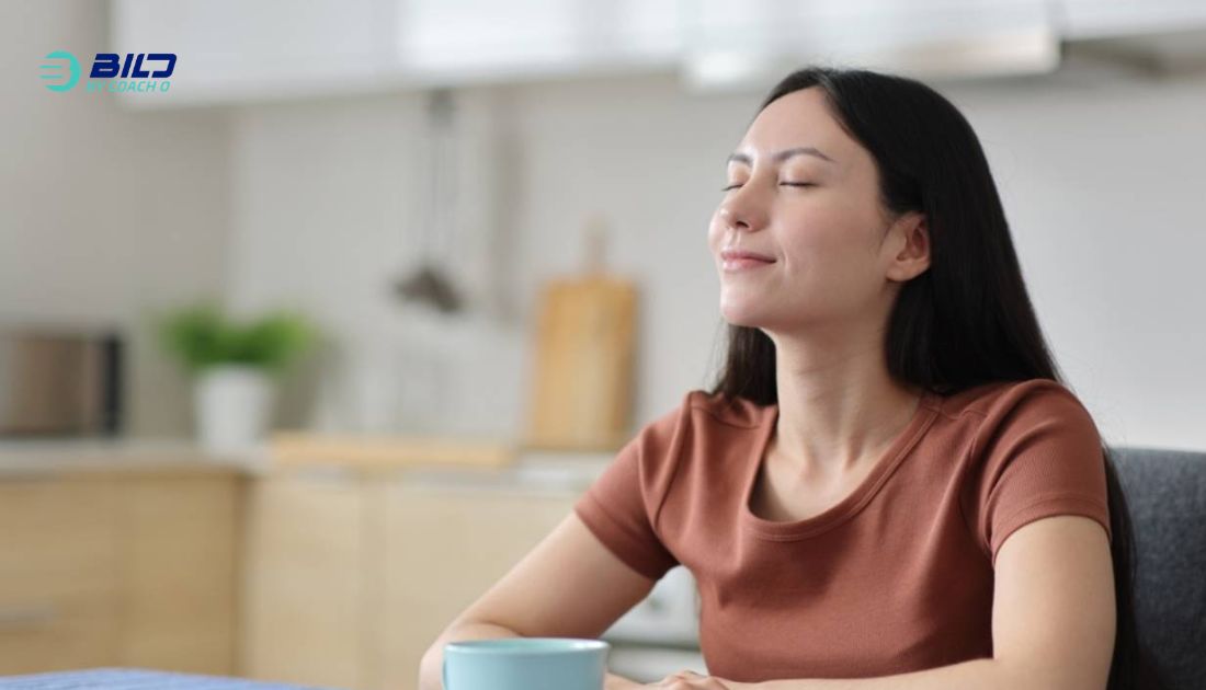 A relaxed woman with closed eyes and a gentle smile sitting in a bright kitchen, enjoying a morning beverage from a blue mug.